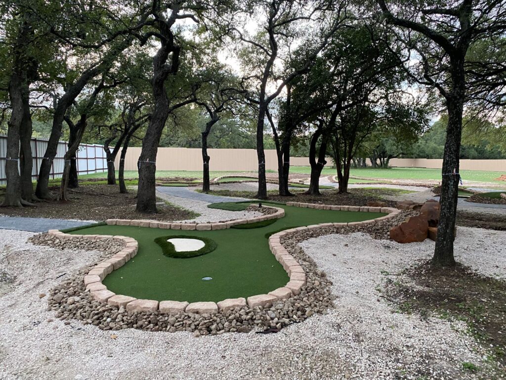 Singular golf hole in a miniature golf course, trees surrounding course, in the woods, sand bordering the hole.