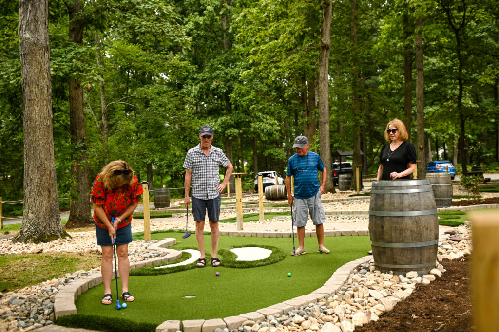 Miniature golf course at a vineyard, people playing on the course, green trees surrounding the course, faux sand bunkers on the hole to emulate a golf course