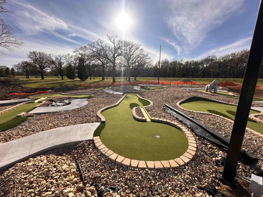 miniature golf hole, sun shining on the course, cement walkways between holes, the golf hole has a shoot-through bridge on it