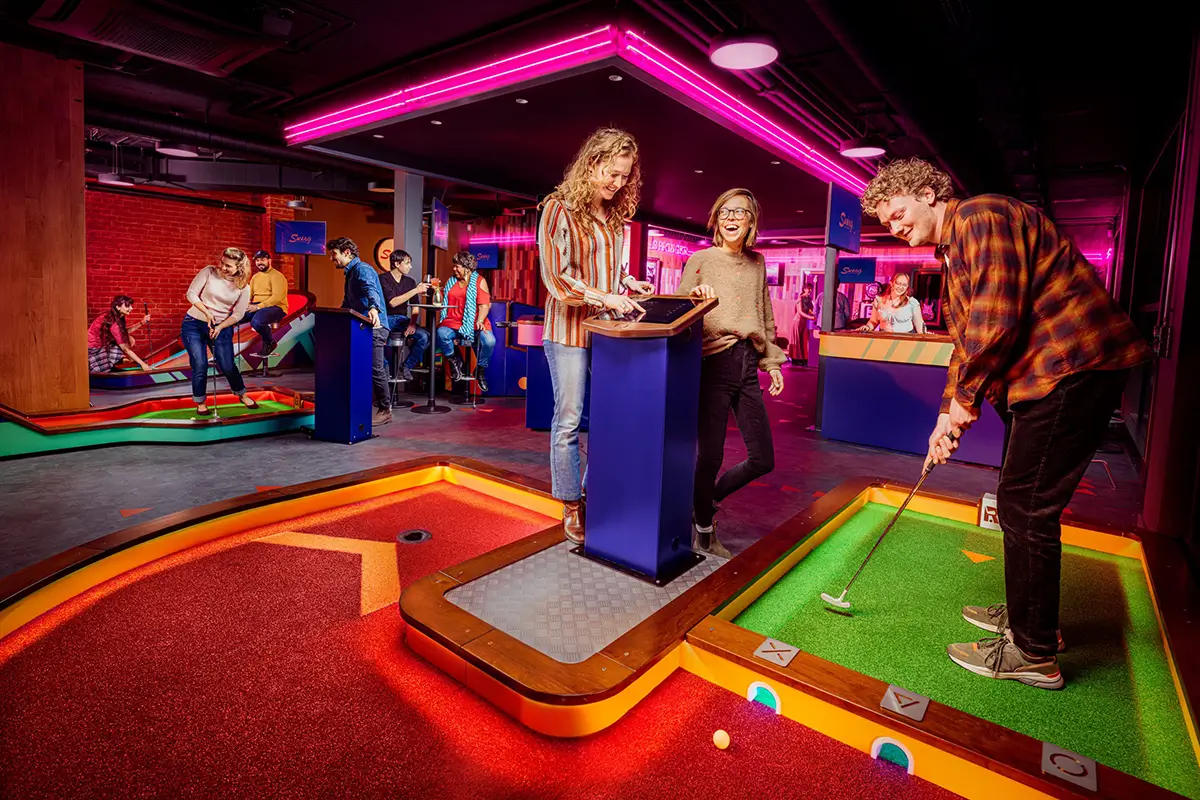 Players putting on RemarkaBall high-tech mini-golf holes as others enter their scores on a kiosk